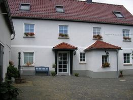 Zweistöckiges Haus mit Blumenfenstern und blauem Sitzplatz, gepflasterter Hof, bewölkter Himmel.