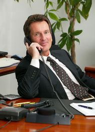 Mann im Anzug telefoniert im Büro, sitzt am Schreibtisch mit Pflanzen im Hintergrund.