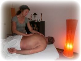 Frau massiert liegenden Mann auf einem Tisch, sanftes Licht von einer Lampenscheint im Raum.