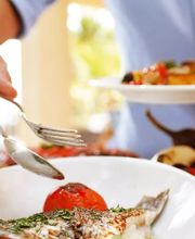 Persona sirviendo pescado con verduras en un plato, rodeado de otros alimentos.