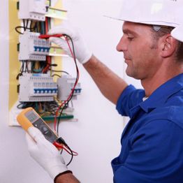 Electrician in blue uniform using a multimeter to check wiring in a circuit breaker panel.