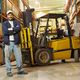 Man in warehouse standing beside a yellow forklift, surrounded by stacked shelves.