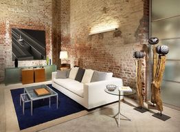 Loft-Wohnzimmer mit Ziegelwänden, weißem Sofa, blauer Teppich und dekorativen Holzskulpturen.