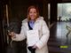 Donna sorridente apre una porta, tenendo dei documenti in mano, indossa un cappotto bianco.