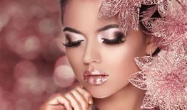 Frau mit glänzendem Makeup und rosa Blumenaccessoire posiert vor einem rosa glitzernden Hintergrund.