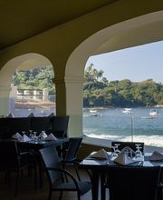 Restaurante con vista al mar, mesas preparadas y paisaje de playa y colinas al fondo.