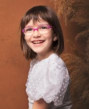 Niña sonriente con gafas rosas y blusa blanca, fondo marrón.