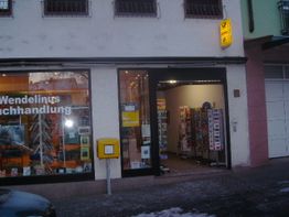 Fassade einer Buchhandlung mit Post-Schild und Zeitschriftenständern am Eingang.