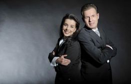 Ein Mann und eine Frau in Anzügen stehen Rücken an Rücken und schauen in die Kamera.