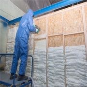 Person in blue suit spraying foam insulation on a wooden wall structure.
