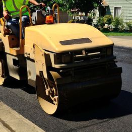 Worker operates a yellow road roller on fresh asphalt near a sidewalk on a sunny day.