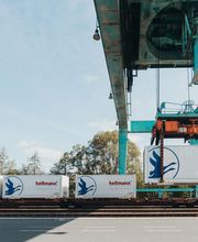 Grúa levantando un contenedor Hellmann en un depósito ferroviario con cielo despejado.