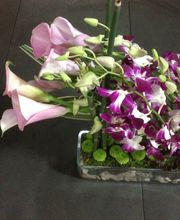 Arreglo floral con lirios rosas y orquídeas moradas en un recipiente rectangular.