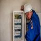 Electrician in blue uniform checking a circuit breaker panel on a wall.