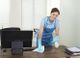 Smiling woman in uniform cleaning a desk in an office with spray and cloth.