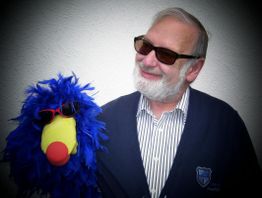 Ein Mann mit Bart lächelt, trägt Sonnenbrille, neben ihm ein blauer Plüschvogel mit Sonnenbrille.