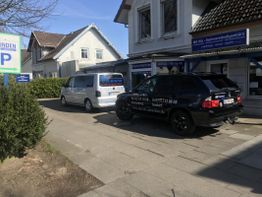 Zwei geparkte Autos vor einem Gebäude, sonniger Tag, blaues Schild im Hintergrund.