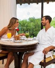 Pareja desayunando en un balcón con vista al bosque, platos, jugos y una rosa en la mesa.