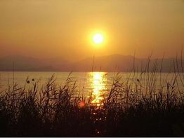 Sonnenuntergang über einem ruhigen See mit Schilf im Vordergrund und Bergen im Hintergrund.