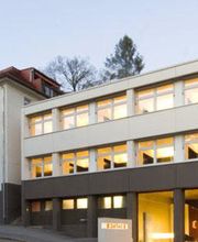 Moderne Gebäudeansicht bei Dämmerung, mit beleuchteten Fenstern und Bäumen im Hintergrund.