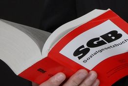 Person hält ein geöffnetes Buch mit rotem Umschlag und „SGB Sozialgesetzbuch“ auf dem Cover.