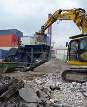 Bagger bei Abrissarbeiten mit Bauschutt im Vordergrund in einer industriellen Umgebung.