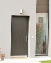 Graue Hausfassade mit brauner Tür, großem Fenster und Pflanzen.