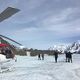 Helicopter on snowy terrain, with people walking toward distant snow-capped mountains.