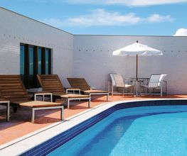 Piscina em terraço com espreguiçadeiras de madeira e guarda-sol branco. Céu azul ao fundo.