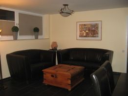 Wohnzimmer mit schwarzen Ledersofas, Holztisch, Kunstwerk an der Wand und dekorativen Pflanzen.