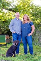 Ein Paar steht mit einem Hund auf einer Wiese, Bäume im Hintergrund, sie tragen blaue Kleidung.