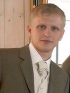 Mann in einem Anzug mit Krawatte, im Hintergrund helle Holzwand.