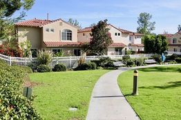 Pathway leading to houses with red-tiled roofs, surrounded by greenery and benches.