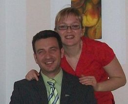 Zwei lächelnde Personen, Mann im Anzug, Frau in rotem Shirt, vor einem Gemälde stehend.