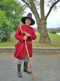Ein Mann in mittelalterlicher Kleidung mit Hut und Stock steht vor einem Baum auf einer Straße.