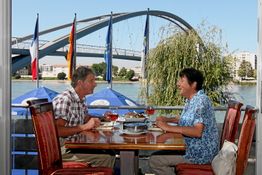 Zwei Personen essen draußen an einem Tisch mit Flussblick, Brücke und wehenden Flaggen im Hintergrund.