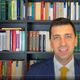 Uomo in giacca e cravatta gialla davanti a una libreria piena di libri colorati.