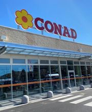 Esterno di un supermercato Conad con insegna rossa e fiore giallo sul tetto.