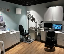 Optometriekamer met oogmeetapparatuur, bureau, stoelen, computer en opslagkasten.