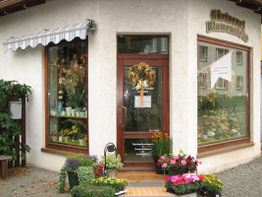 Blumenladen-Eingang mit Pflanzen und Blumen davor, dekoriert mit einem Kranz an der Tür.