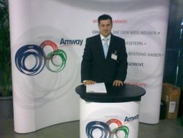 Mann im Anzug am Amway-Stand mit Grafiken und Text auf einem Roll-up im Hintergrund.