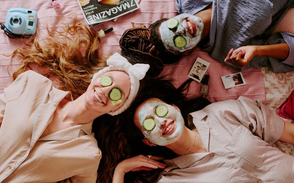 Drei Personen entspannen mit Gesichtsmasken und Gurkenscheiben auf den Augen gemeinsam auf einem Bett.