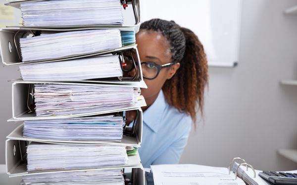 Mitarbeiterin im Büro hinter einem hohen Stapel mit Aktenordnern und Dokumenten.
