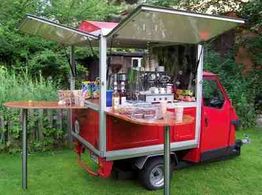 Roter mobiler Kaffeewagen mit Klapptischen, Kaffeemaschine und Zubehör auf grünem Rasen.