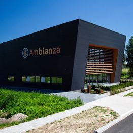 Modern gebouw met het logo van Ambianza, omgeven door groen en een heldere blauwe lucht.