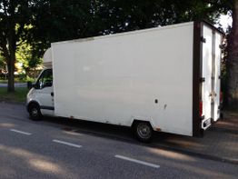 Weißer Lieferwagen parkt am Straßenrand vor einem Park mit Bäumen im Hintergrund.