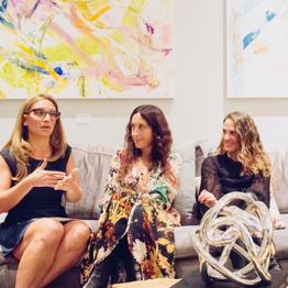 Three women sitting on a couch in discussion, with colorful abstract art in the background.