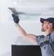 Worker in a cap and gloves smoothing ceiling drywall with a trowel tool.