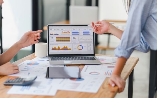 Zwei Kollegen einer Firma besprechen, die Performance Zahlen. Zwischen ihnen steht ein Laptop. Auf dem Display sind verschiedene Diagramme zu sehen.