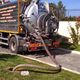 Camion autospurghi in azione, con tubo collegato a tombino in un vialetto residenziale.